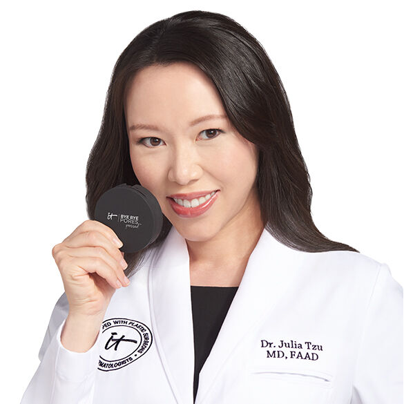 A photo of IT Cosmetics advisor Dr. Julia Tzu wearing a white doctor's coat with an embroidered IT Cosmetics seal, holding a black compact in her hand.