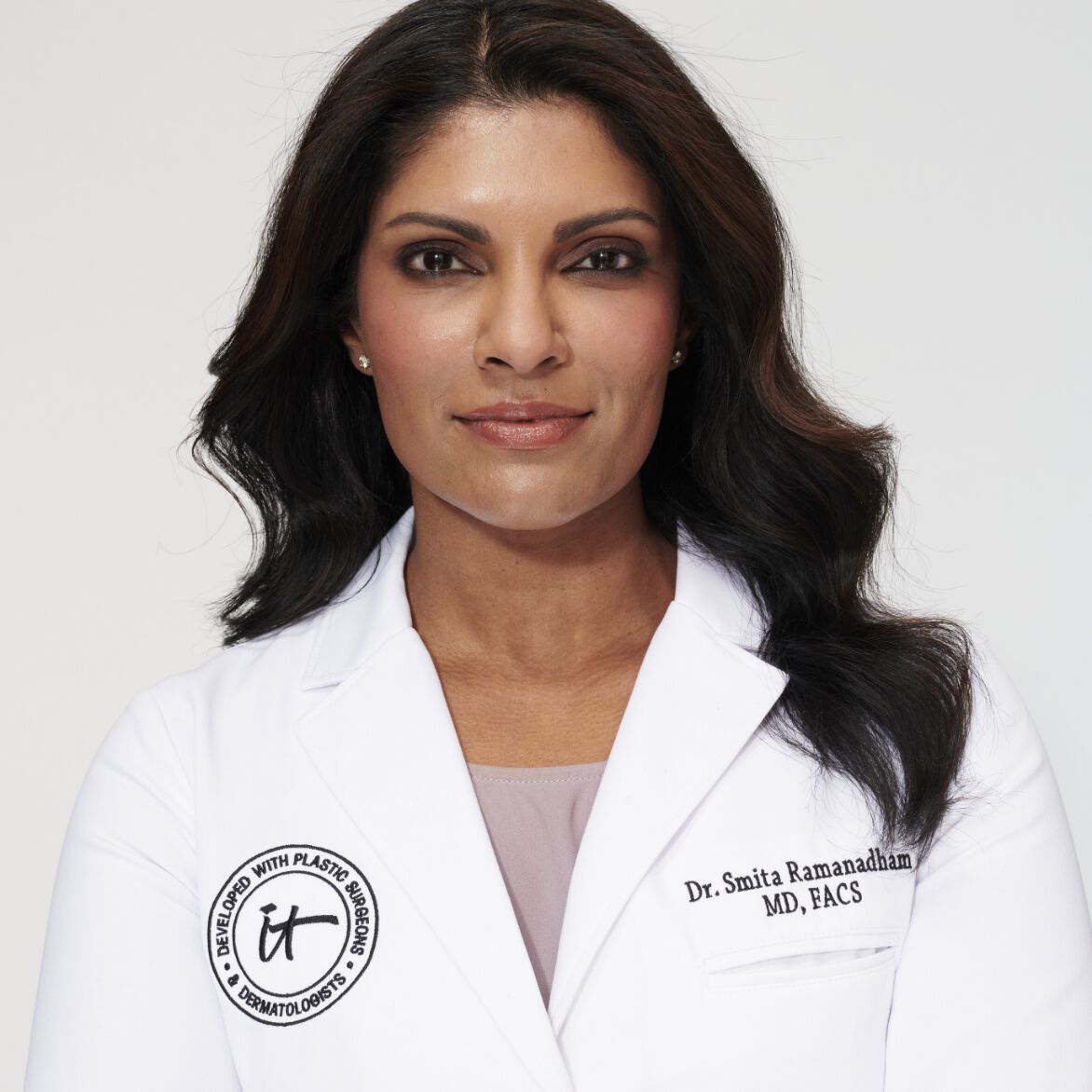A headshot of IT Cosmetics advisor Dr. Smita R. Ramanadham wearing a white doctor's coat with an embroidered IT Cosmetics seal.