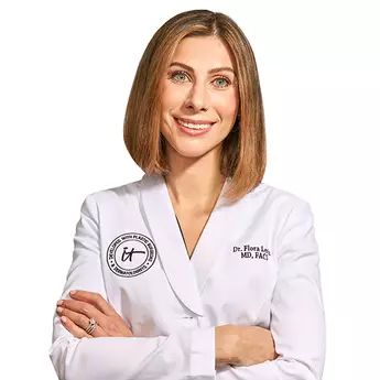 A headshot of IT Cosmetics advisor Dr. Flora Levin wearing a white doctor's coat with an embroidered IT Cosmetics seal.