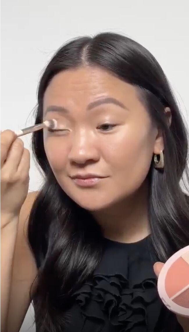 A photo of a woman with long dark brown hair holding an eye shadow pallet and applying eyeshadow on her eyelid with a brush.
