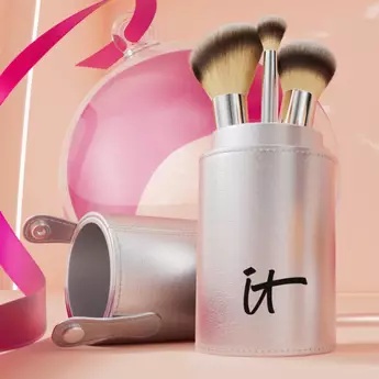A photo of a silver makeup brush holder with "IT" imprinted on the container holding three makeup brushes on a pink background.