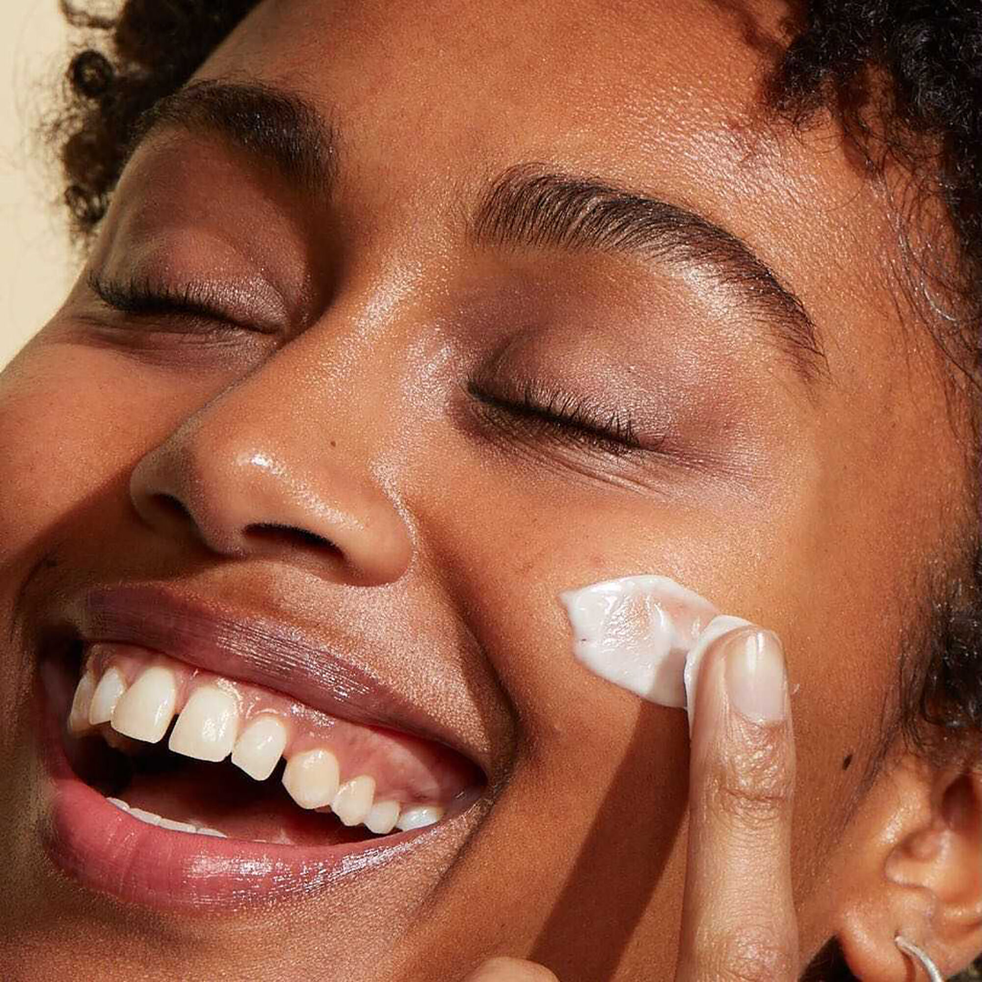 A photo of a woman smiling while rubbing a facial cream into her skin.