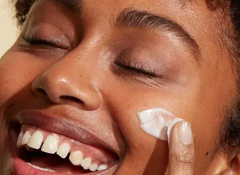 A photo of a woman smiling while rubbing a facial cream into her skin.