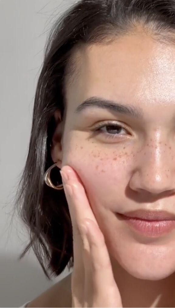 A close-up photo of half a woman's face as she applies a product to her cheek.