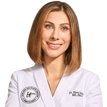 A headshot of IT Cosmetics advisor Dr. Flora Levin wearing a white doctor's coat with an embroidered IT Cosmetics seal.