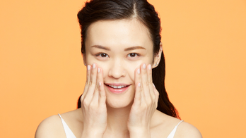 A photo of a woman applying face cream with both hands and an orange background