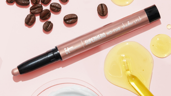 Product shot of IT Cosmetics eyeshadow stick surrounded by coffee beans and a yellow serum