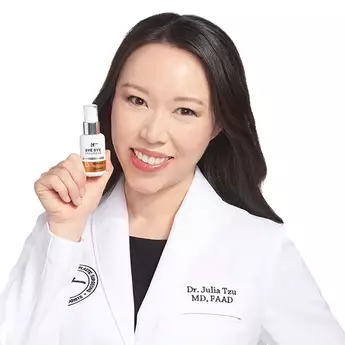 A photo of IT Cosmetics advisor Dr. Julia Tzu wearing a white doctor's coat with an embroidered IT Cosmetics seal, holding a serum bottle in her hand.