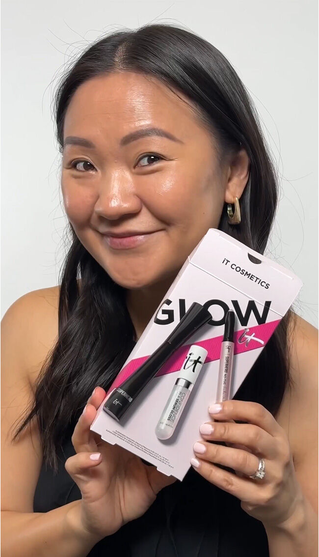 A chest-up photo of a woman with long dark brown hair smiling and looking at the camera, holding the IT Cosmetics Glow IT gift box.