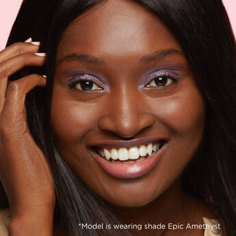 A closely cropped photo of a model with deep skin's face smiling at the camera, with text that reads "*Model is wearing shade epic Amethyst."