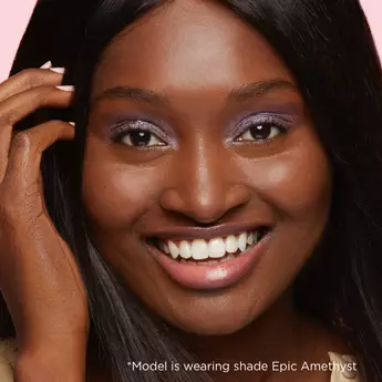 A closely cropped photo of a model with deep skin's face smiling at the camera, with text that reads "*Model is wearing shade epic Amethyst."