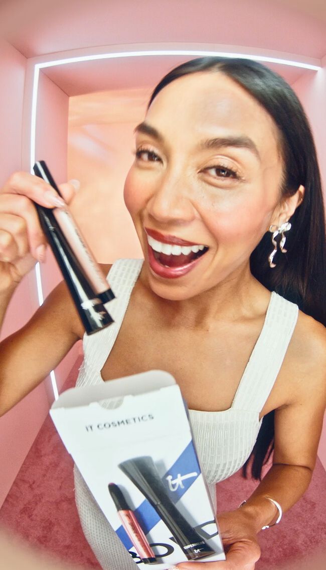 A photo of a woman with long dark brown hair opening an IT Cosmetics Celebrate Bold Eyes Eyeshadow Stick & Mascara Gift Set box and pulling out the products.