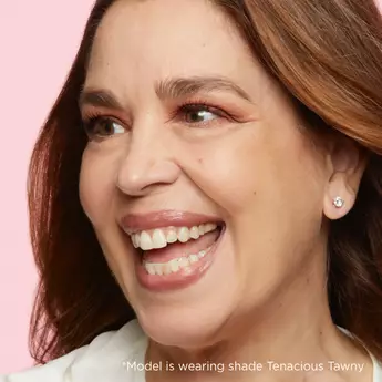 A close-up photo of a woman smiling and looking off to the side with text that reads, "*Model is wearing shade Tenacious Tawny."