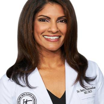 A headshot of IT Cosmetics advisor Dr. Smita R. Ramanadham wearing a white doctor's coat with an embroidered IT Cosmetics seal.