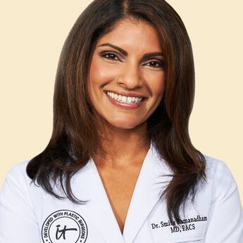 A headshot of IT Cosmetics advisor Dr. Smita R. Ramanadham wearing a white doctor's coat with an embroidered IT Cosmetics seal.
