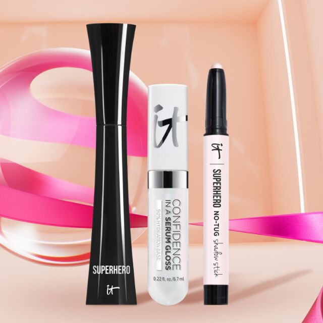 Three IT Cosmetics products, Superhero IT Mascara, Confidence In A Serum Gloss, Superhero No Tug Shadowstick, and a multicolored pink background.