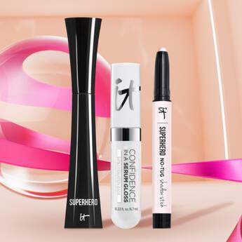 Three IT Cosmetics products, Superhero IT Mascara, Confidence In A Serum Gloss, Superhero No Tug Shadowstick, and a multicolored pink background.